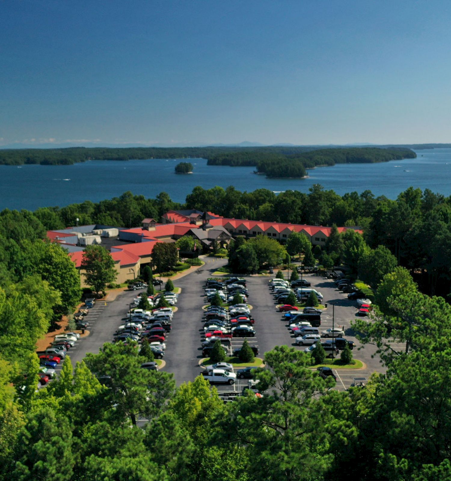 Aerial view of a large building with red roofs, surrounded by trees, near a lake with parked cars.